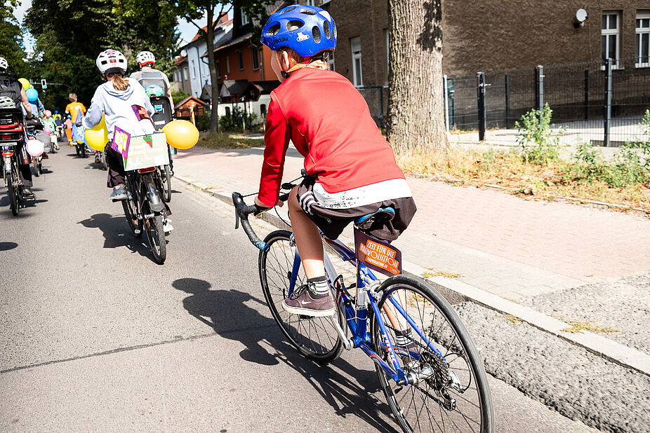 Teilnehmende der Kidical-Mass-Fahrrad-Demo in Hohen Neuendorf im September 2023. Ein von schräg hinten fotografiertes Kind mit Fahrradhelm fährt auf einem Fahrrad und nimmt an einer Fahrrad-Demo teil.